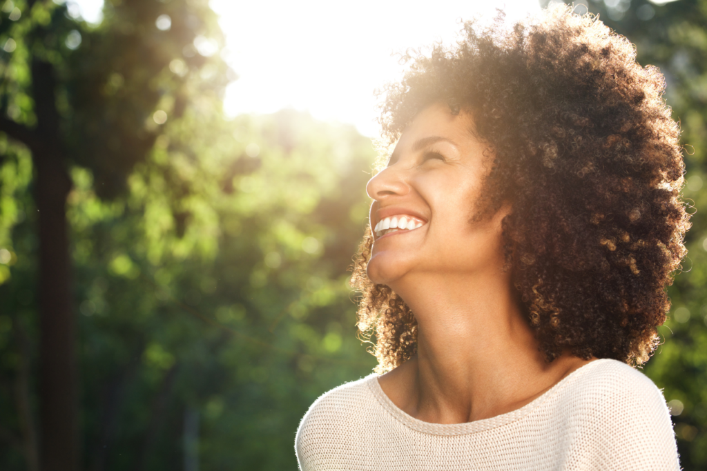 Femme souriante aux cheveux bouclés, regard levé vers la lumière du soleil, en extérieur dans un environnement naturel verdoyant.