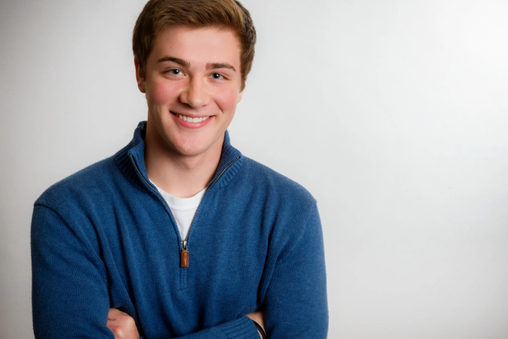 Jeune personne souriante, cheveux courts châtain clair, portant un pull bleu à col zippé sur fond clair. Bras croisés, posture détendue, expression ouverte et sympathique.