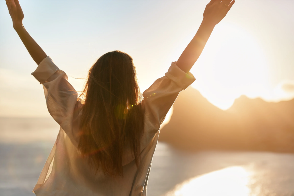 Image d'une femme de dos, les bras levés et écartés comme signe de renouveau face au lever de soleil sur le mer, synonyme du renouveau apporté grâce aux séances de sophrologie à Lyon et Villeurbanne